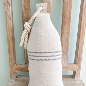 May include: A cream-colored doorstop with a rope handle and two blue stripes. The doorstop is sitting on a wooden chair.