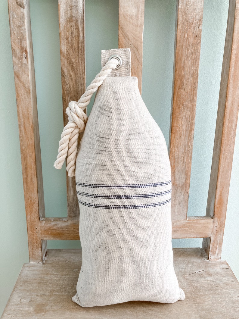 May include: A cream-colored doorstop with a rope handle and two blue stripes. The doorstop is sitting on a wooden chair.