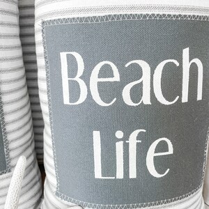 May include: Two white and gray striped throw pillows with a gray square patch on the front. The patch has the text "Beach Life" in white lettering.