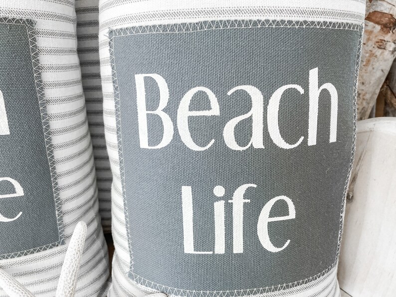 May include: Two white and gray striped throw pillows with a gray square patch on the front. The patch has the text "Beach Life" in white lettering.