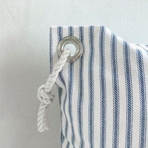 May include: A white canvas bag with blue stripes and a white rope drawstring. The bag has a metal grommet with a white rope threaded through it.