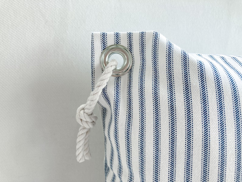 May include: A white canvas bag with blue stripes and a white rope drawstring. The bag has a metal grommet with a white rope threaded through it.
