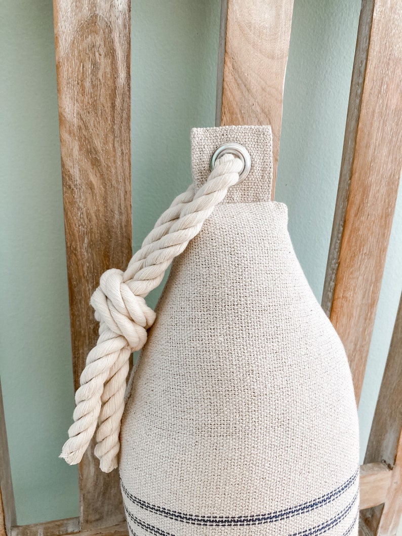 May include: A beige doorstop with a rope handle and blue stripes. The doorstop has a metal ring on the top.