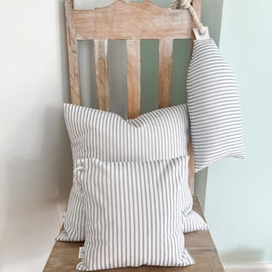 May include: Three striped pillows in white and blue on a wooden chair. The pillows are arranged in a stack, with the largest pillow at the bottom and the smallest pillow at the top. The chair is made of light wood and has a simple design.