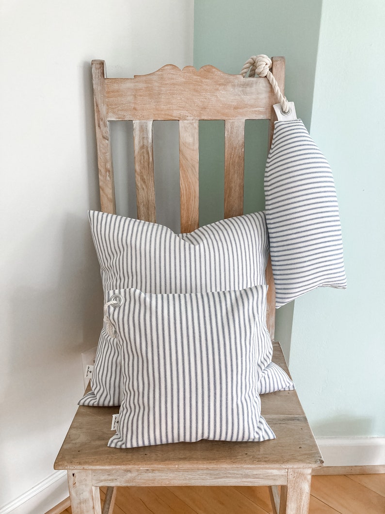 May include: Three striped pillows in white and blue on a wooden chair. The pillows are arranged in a stack, with the largest pillow at the bottom and the smallest pillow at the top. The chair is made of light wood and has a simple design.