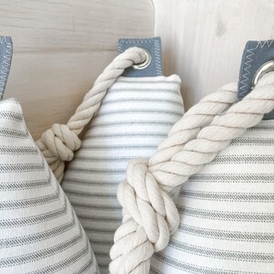 May include: Three white and gray striped fabric doorstops with a thick white rope tied around them. The doorstops have a gray fabric loop with a metal ring.