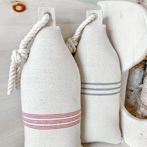 May include: Two cream-colored fabric pillows with rope handles. One pillow has red stripes and the other has blue stripes.