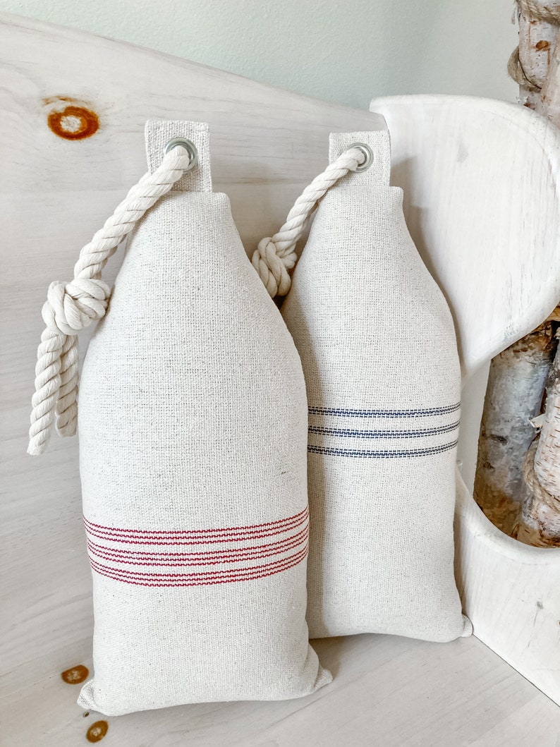 May include: Two cream-colored fabric pillows with rope handles. One pillow has red stripes and the other has blue stripes.