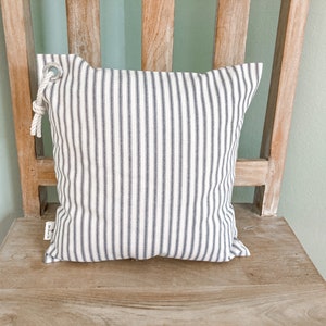 May include: A white and gray striped pillow with a rope loop and a metal ring. The pillow is sitting on a wooden chair.