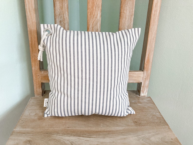 May include: A white and gray striped pillow with a rope loop and a metal ring. The pillow is sitting on a wooden chair.