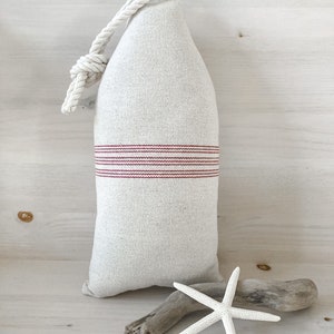 May include: A white linen pillow with red stripes and a rope handle. The pillow is shaped like a buoy and is sitting on a white wooden surface.