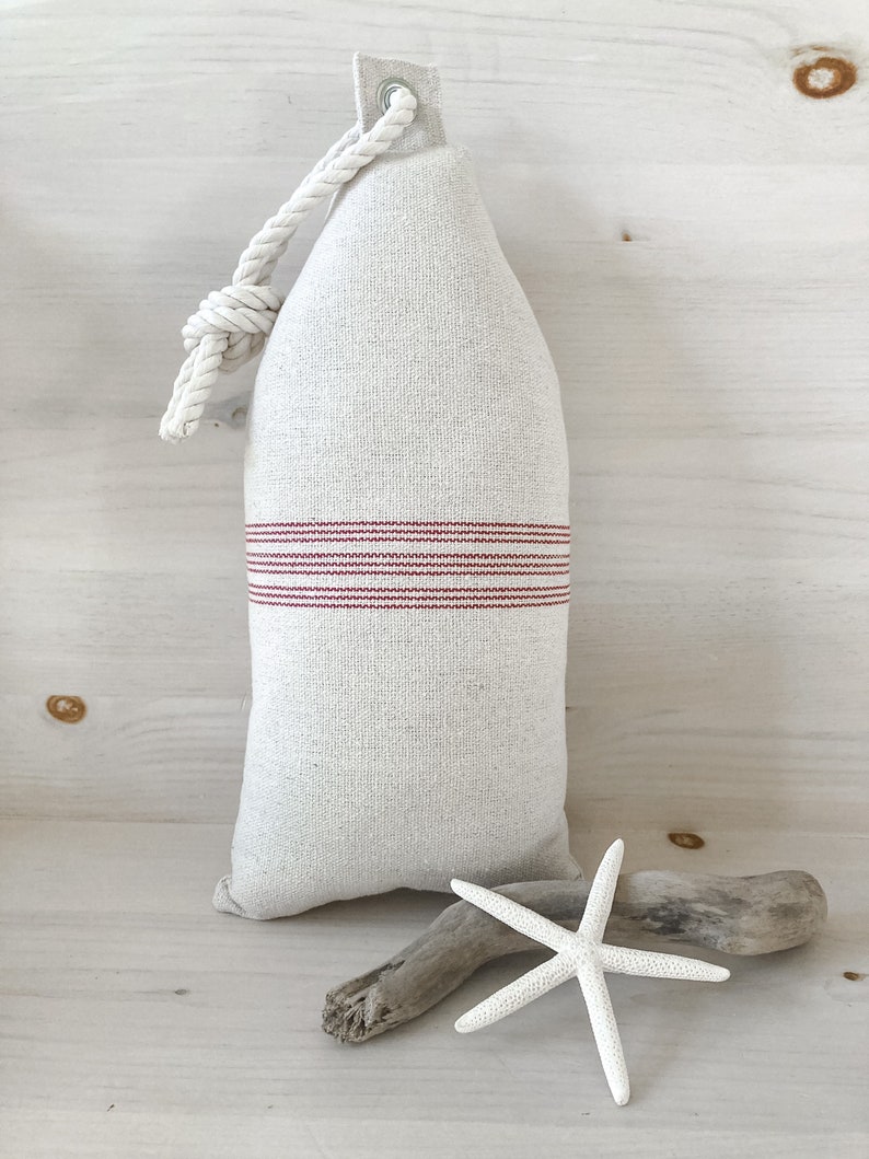 May include: A white linen pillow with red stripes and a rope handle. The pillow is shaped like a buoy and is sitting on a white wooden surface.
