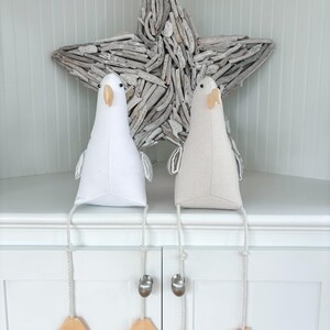 May include: Two decorative bird-shaped fabric ornaments, one white and one beige, with dangling legs. Each bird has a beak, eyes, and small wings. The legs are attached to a rope with a metal weight and a wooden foot. A driftwood star is in the background.