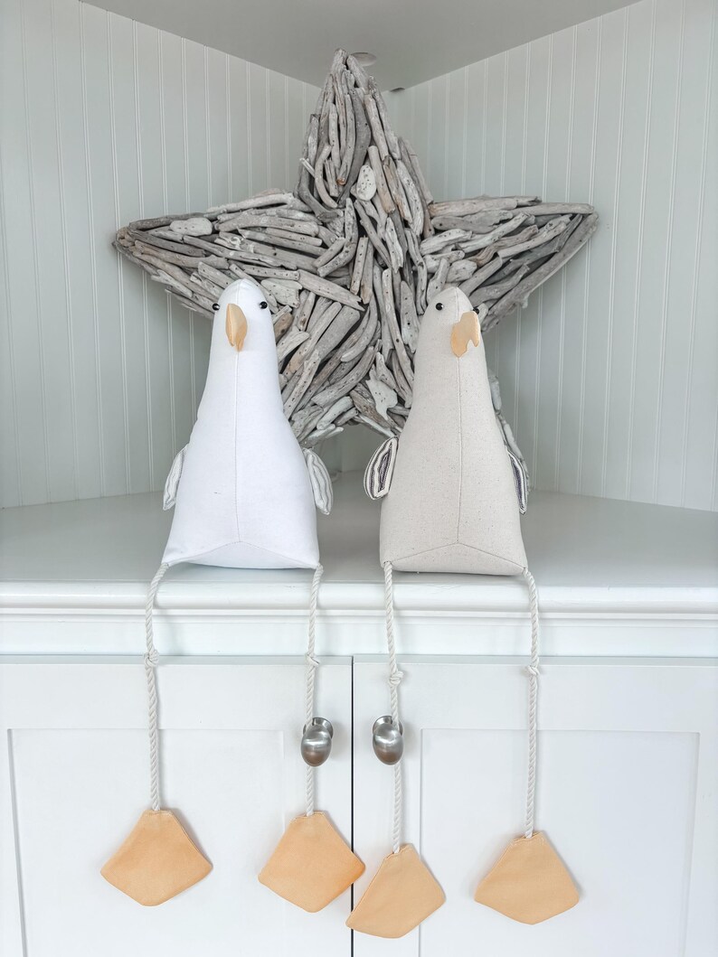 May include: Two decorative bird-shaped fabric ornaments, one white and one beige, with dangling legs. Each bird has a beak, eyes, and small wings. The legs are attached to a rope with a metal weight and a wooden foot. A driftwood star is in the background.