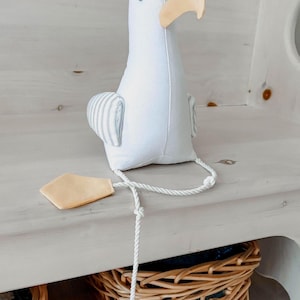 May include: A white stuffed seagull toy with a light yellow beak and feet. The seagull has small black eyes and gray and white striped wings. A white rope is attached to the feet.