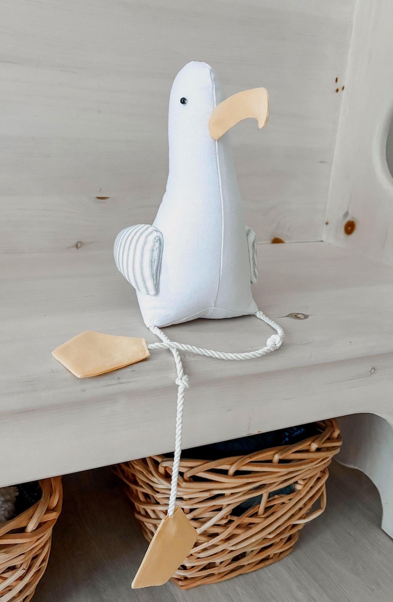 May include: A white stuffed seagull toy with a light yellow beak and feet. The seagull has small black eyes and gray and white striped wings. A white rope is attached to the feet.