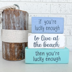 Puede incluir: Tres bloques de madera apilados uno encima del otro. El bloque superior es azul claro y dice "if you're lucky enough". El bloque del medio es blanco y dice "to live at the beach". El bloque inferior es turquesa y dice "then you're lucky enough".
