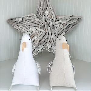 May include: Two plush seagull figures with white and beige bodies, black eyes, and tan beaks. They have small wings and are positioned in front of a large driftwood star. The seagulls have rope legs, and the background is a white cabinet.