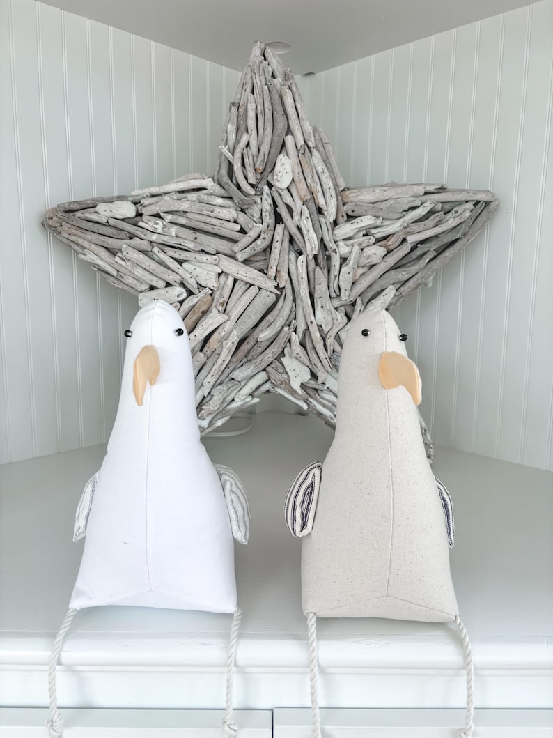 May include: Two plush seagull figures with white and beige bodies, black eyes, and tan beaks. They have small wings and are positioned in front of a large driftwood star. The seagulls have rope legs, and the background is a white cabinet.