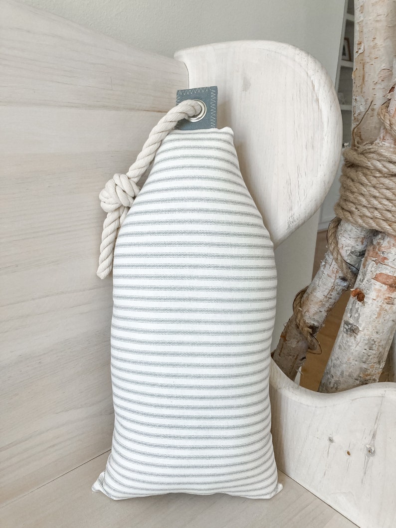 May include: A white and gray striped pillow with a rope loop and a gray fabric tab. The pillow is hanging on a white wooden surface.