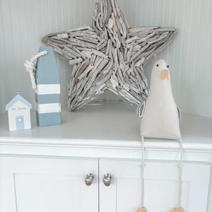 May include: Coastal-themed decor featuring a large driftwood star, a blue and white buoy with rope, a small house with "Sea Breeze" text, and a plush seagull with dangling legs and wooden feet. The items are arranged on a white cabinet.