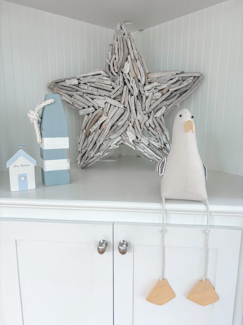 May include: Coastal-themed decor featuring a large driftwood star, a blue and white buoy with rope, a small house with "Sea Breeze" text, and a plush seagull with dangling legs and wooden feet. The items are arranged on a white cabinet.
