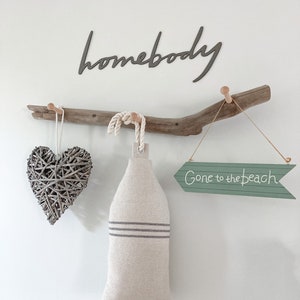 May include: A driftwood branch with three hooks, a woven heart, a linen bag, and a green arrow sign that reads "Gone to the beach". The word "homebody" is written in black on the wall above the branch.