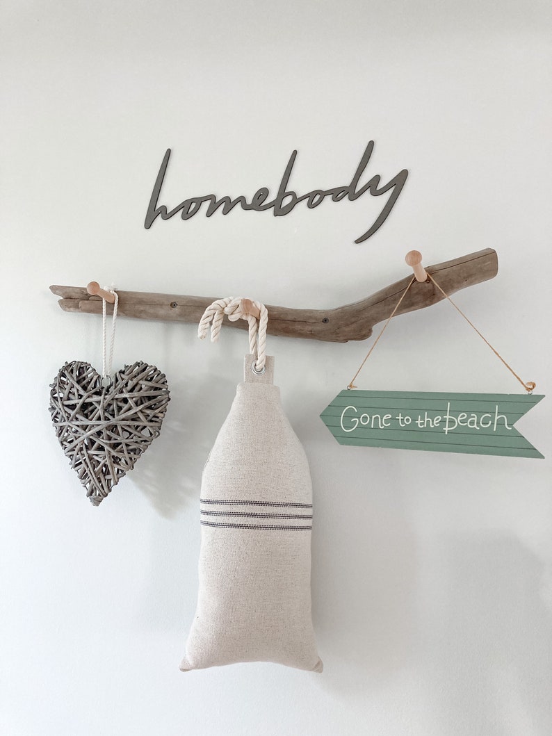 May include: A driftwood branch with three hooks, a woven heart, a linen bag, and a green arrow sign that reads "Gone to the beach". The word "homebody" is written in black on the wall above the branch.