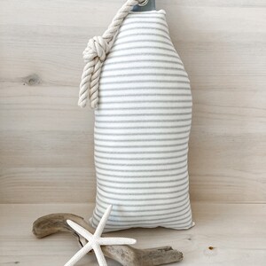 May include: A white and gray striped fabric doorstop with a white rope handle. The doorstop is shaped like a bottle and is sitting on a wooden surface.