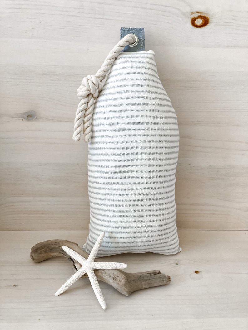 May include: A white and gray striped fabric doorstop with a white rope handle. The doorstop is shaped like a bottle and is sitting on a wooden surface.