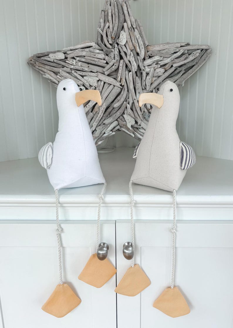 May include: Two seagull-shaped decorative items. One is white, the other is light gray. Both have beige beaks and feet, and striped wings. They hang from a white cabinet with silver knobs, with a driftwood star in the background.