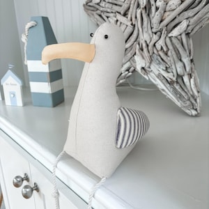 May include: A stuffed seagull with a beige body, a tan beak, and a striped wing. The seagull is sitting on a white surface. Decorative nautical items are in the background, including a blue and white buoy and a driftwood star.