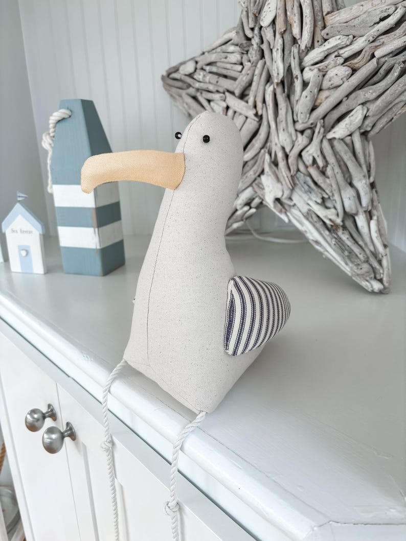 May include: A stuffed seagull with a beige body, a tan beak, and a striped wing. The seagull is sitting on a white surface. Decorative nautical items are in the background, including a blue and white buoy and a driftwood star.