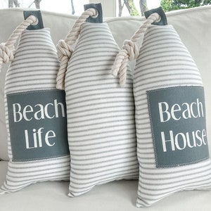 May include: Three white and gray striped doorstops with rope handles. The doorstops have gray fabric squares with white text that reads "Beach Life" and "Beach House".