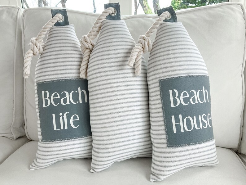May include: Three white and gray striped doorstops with rope handles. The doorstops have gray fabric squares with white text that reads "Beach Life" and "Beach House".