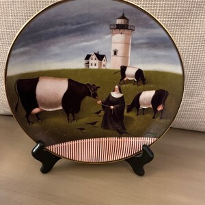 Puede incluir: Un plato redondo de porcelana con una escena pintada de una monja caminando con vacas y un faro en el fondo. El plato está en un soporte negro.