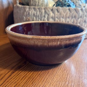 Puede incluir: Un cuenco de cerámica marrón con un esmalte de dos tonos. El interior es de un borgoña intenso, que pasa a un borde beige más claro. El cuenco está sobre una superficie de madera, con una cesta de hilo en el fondo.
