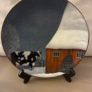 Puede incluir: Una vaca negra y blanca se encuentra en un campo nevado, mirando un granero rojo con un árbol de Navidad en frente. La escena está pintada en un plato redondo de cerámica blanca con un borde dorado.