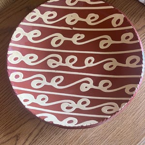 Può includere: Un piatto rotondo color terracotta con un motivo a onde color crema. Il design del piatto presenta linee orizzontali di volute collegate, creando un aspetto decorativo e artistico. Il piatto è probabilmente in ceramica o terracotta.