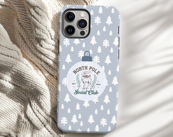 May include: A phone case with a light blue background and a pattern of white Christmas trees. The case features a circular design with the text "North Pole Social Club" and a reindeer illustration.