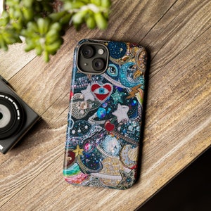 May include: A colourful phone case featuring a variety of embellishments, including stars, hearts, and a cherry. The case has a blue, black, and white background with a red heart in the centre. The case has a camera lens cutout.