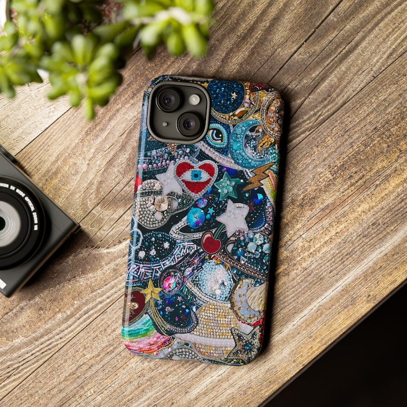 May include: A colourful phone case featuring a variety of embellishments, including stars, hearts, and a cherry. The case has a blue, black, and white background with a red heart in the centre. The case has a camera lens cutout.