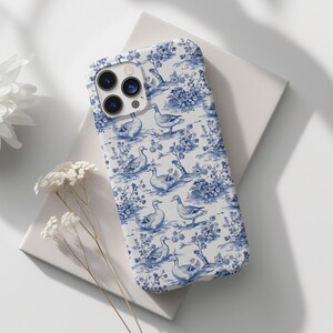 Pu&ograve; includere: Una custodia per iPhone bianca con un motivo blu e bianco di anatre e fiori. La custodia ha un ritaglio per l'obiettivo della fotocamera trasparente.