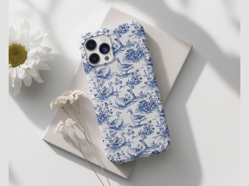 Pu&ograve; includere: Una custodia per iPhone bianca con un motivo blu e bianco di anatre e fiori. La custodia ha un ritaglio per l'obiettivo della fotocamera trasparente.