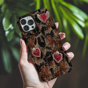 Puede incluir: Una funda de teléfono con estampado de leopardo adornada con tres adornos de corazón rojo. Un corazón tiene alas. La funda se sostiene en una mano, mostrando el diseño y la textura. La funda tiene un recorte para la cámara.
