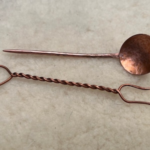May include: Two small copper utensils, a spoon and a fork, on a white fuzzy surface. The spoon has a round bowl and a straight handle. The fork has a twisted handle and two prongs.