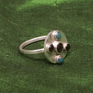 May include: A silver ring with a round center piece featuring three dark red stones and two turquoise stones.