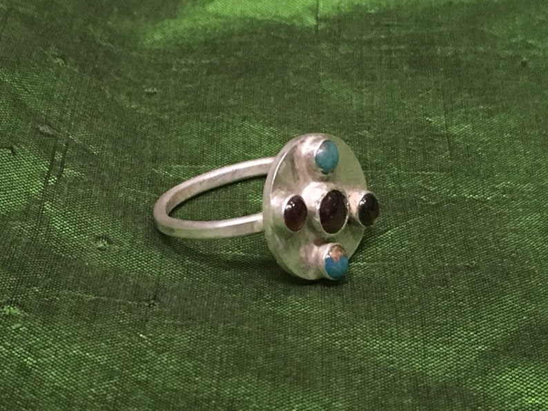 May include: A silver ring with a round center piece featuring three dark red stones and two turquoise stones.