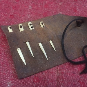 May include: A brown leather case with a black cord closure holds a set of six bone sewing needles. The needles are of varying lengths and have a pointed tip.