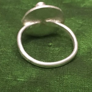May include: A silver ring with a round, flat top. The ring is on a green fabric background.